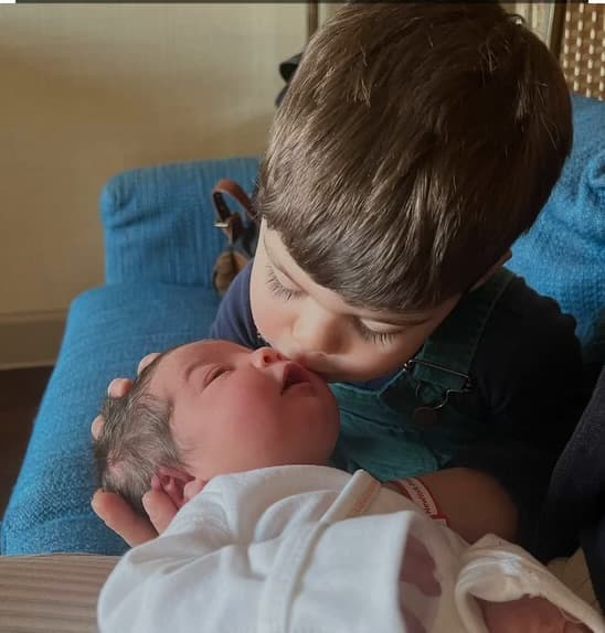 A young boy in green overalls leans down to kiss a newborn baby's cheek.